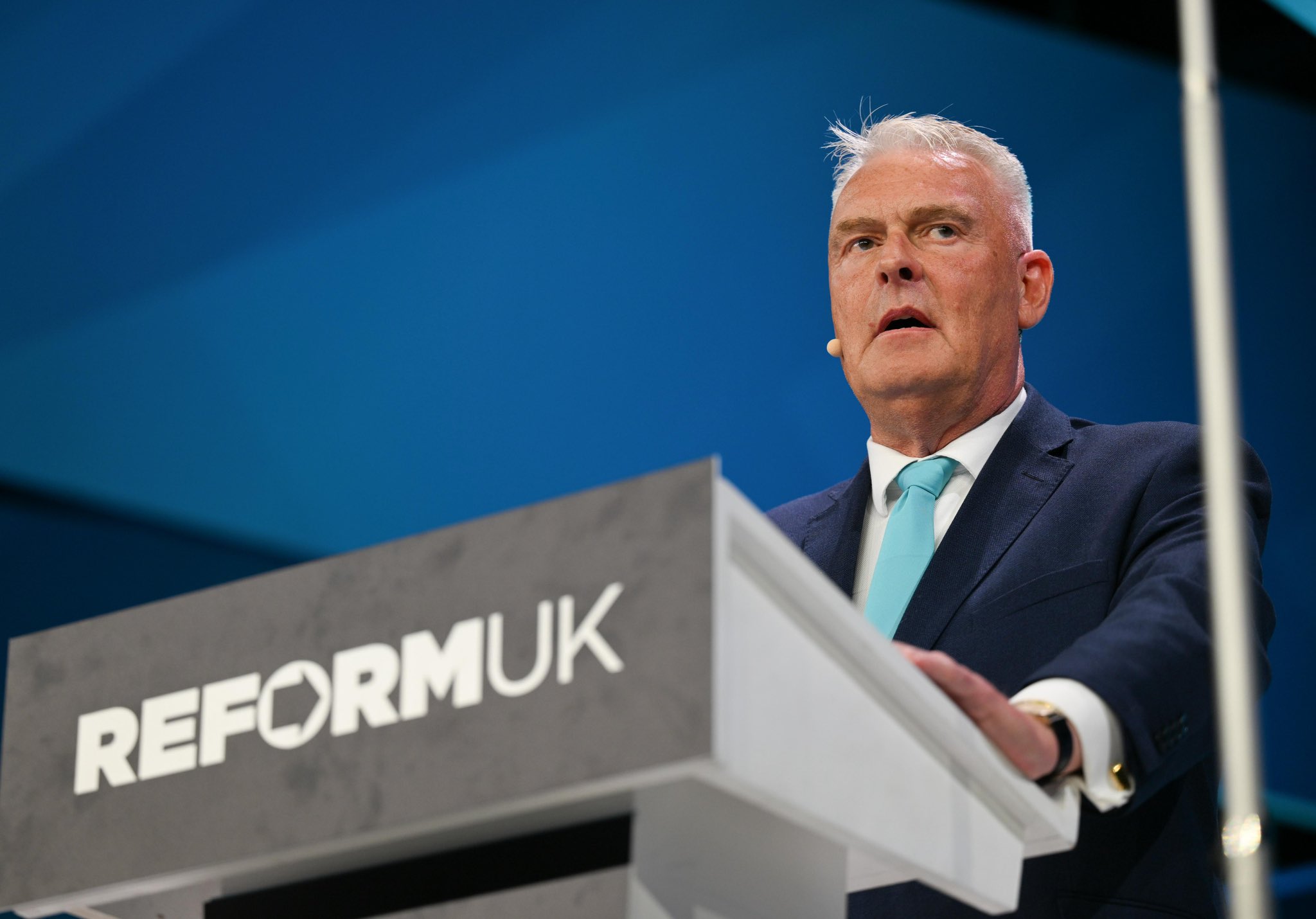 Lee Anderson opens Reform UK Conference 2026 from the podium in Birmingham, wearing a navy suit and turquoise tie beneath Reform UK branding.