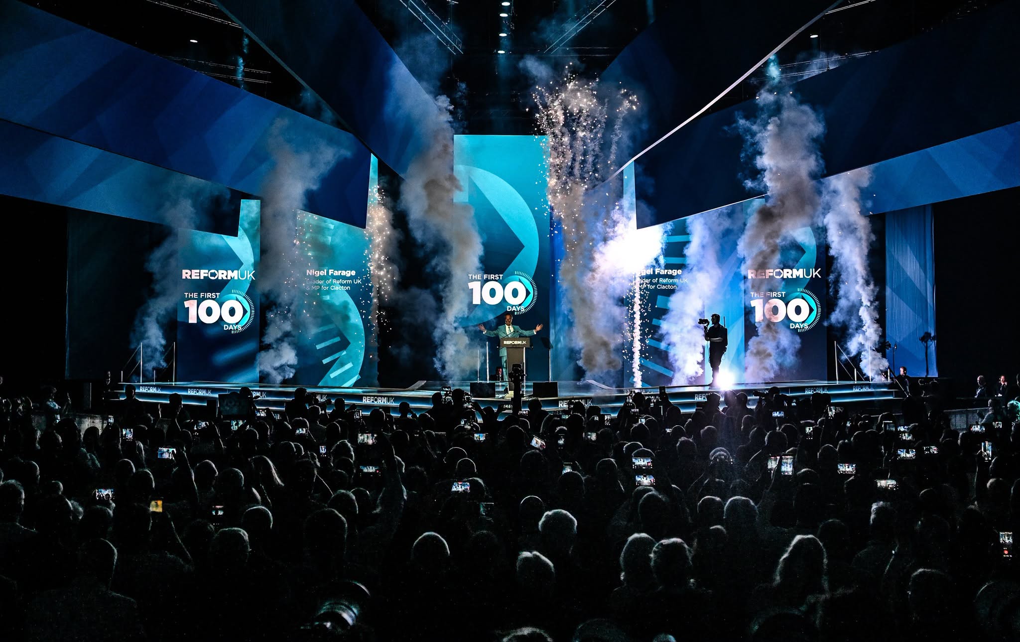 Nigel Farage opens his arms on the Reform UK Conference 2026 main stage in Birmingham as pyrotechnics erupt beneath The First 100 Days branding and delegates film from the hall.
