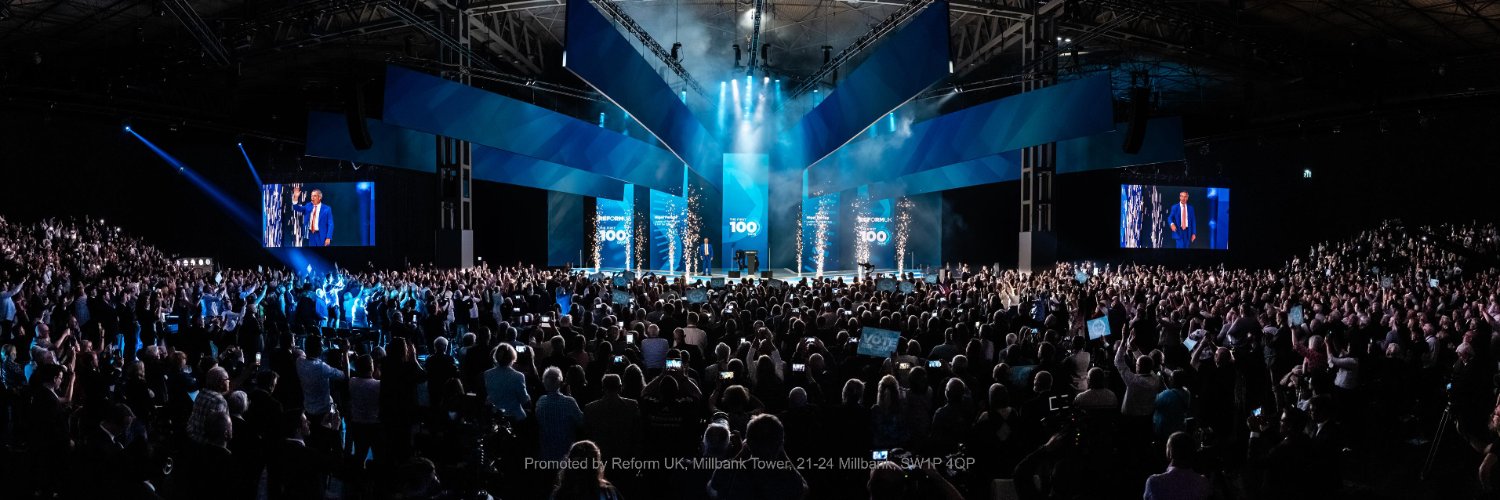 Reform UK Conference 2026 at the Birmingham NEC: a packed hall faces the stage under The First 100 Days banner.