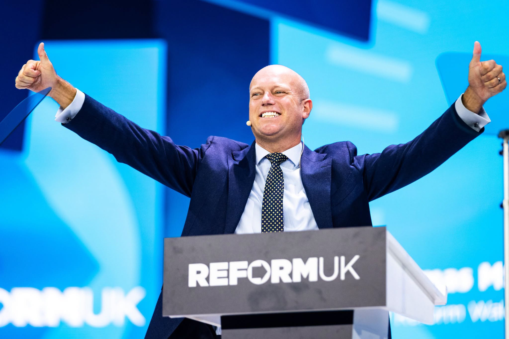 Dan Thomas speaks at Reform UK Conference 2026 in Birmingham, giving a thumbs-up from the Reform UK podium in a navy suit.