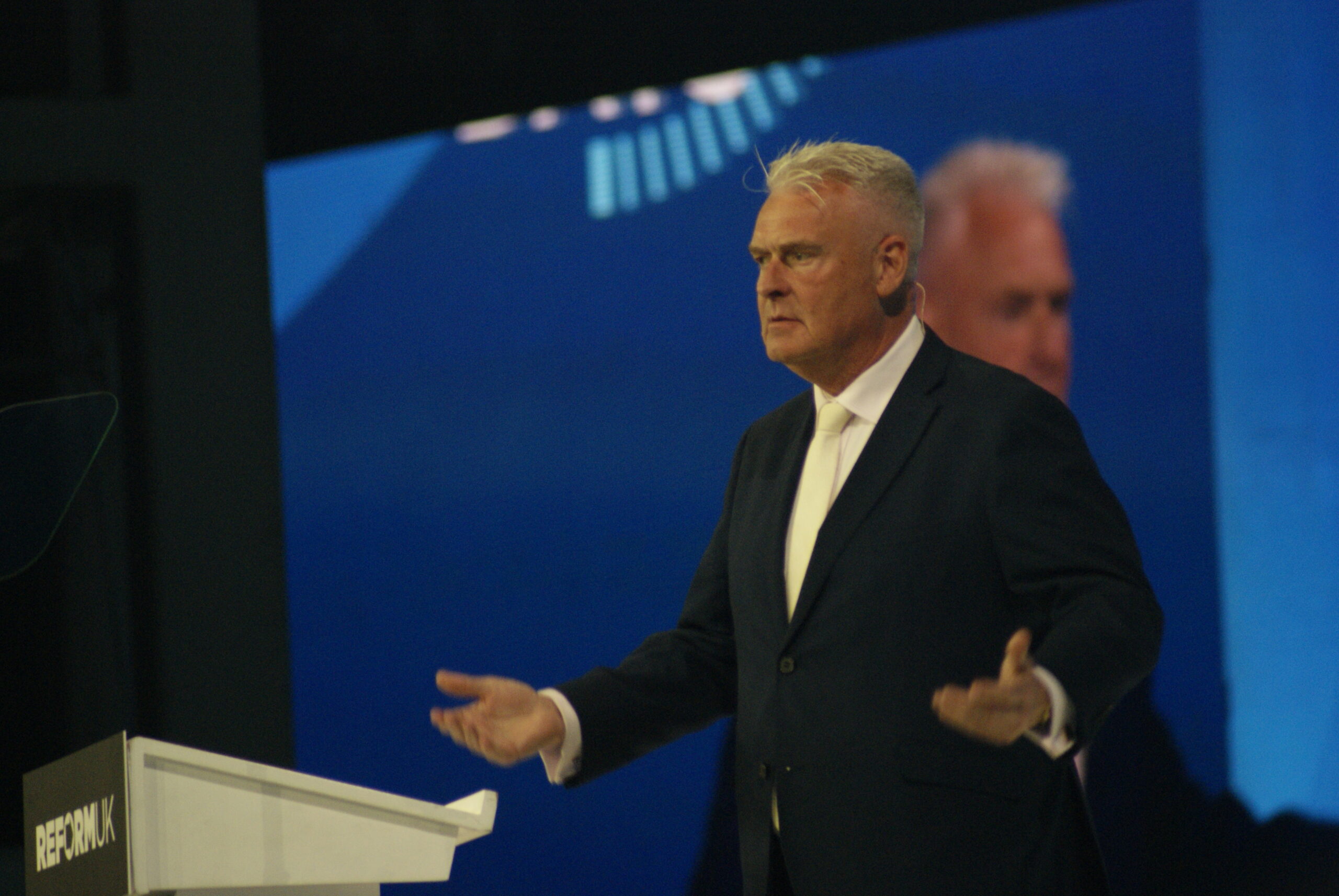 Lee Anderson welcomes delegates back to Reform UK Conference 2026 on Saturday, gesturing from the Reform UK podium in a navy suit.