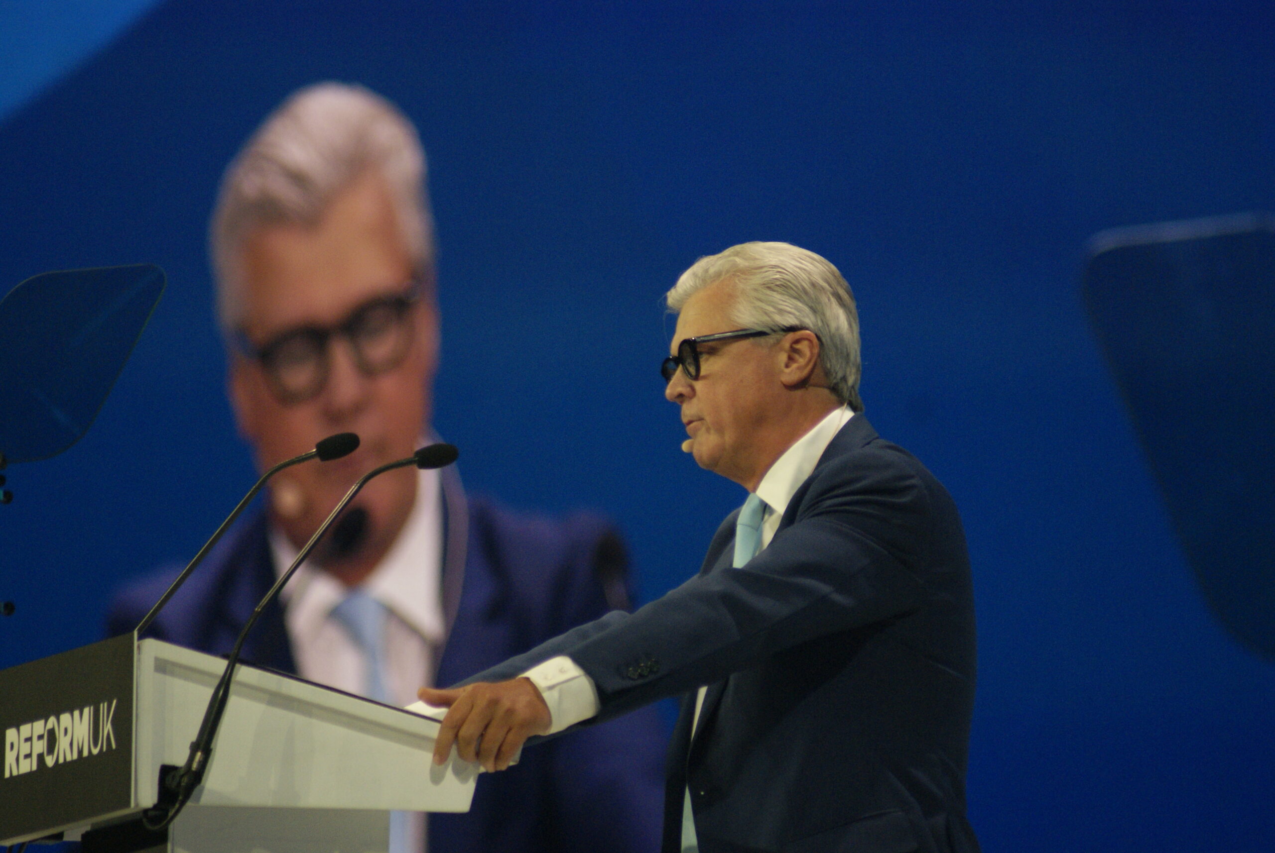 Malcolm Offord speaks from a Reform UK podium at conference, with a large screen behind him showing his portrait against a blue backdrop.