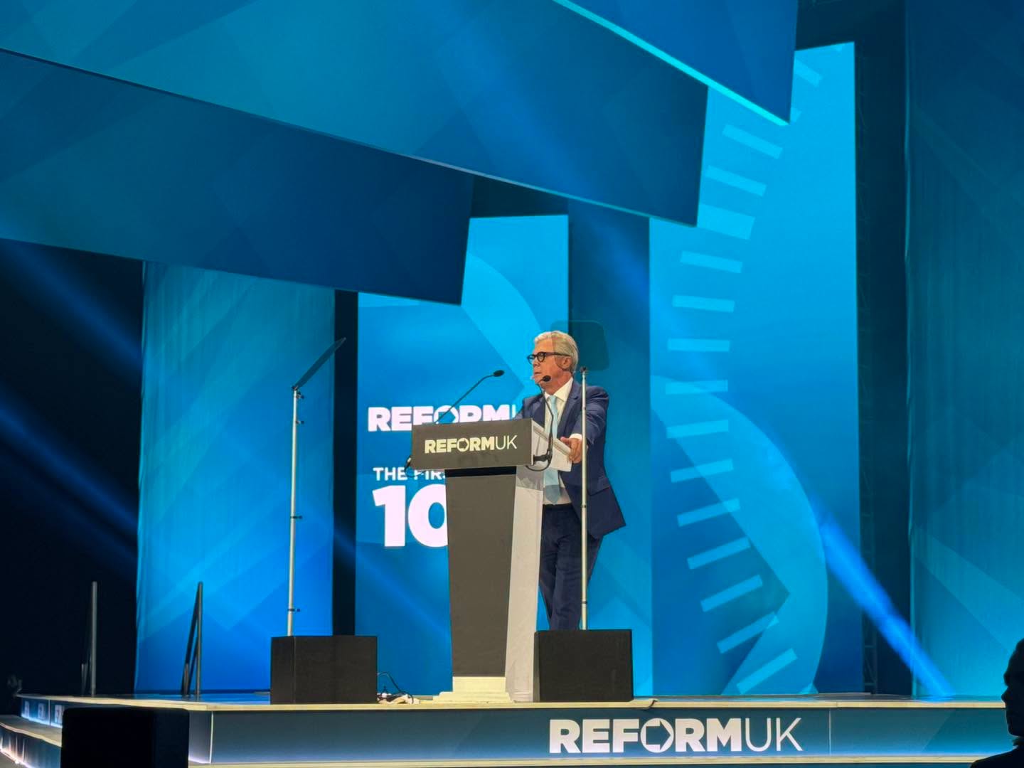 Malcolm Offord speaks at the Reform UK Conference 2026 podium beneath The First 100 Days branding in Birmingham.
