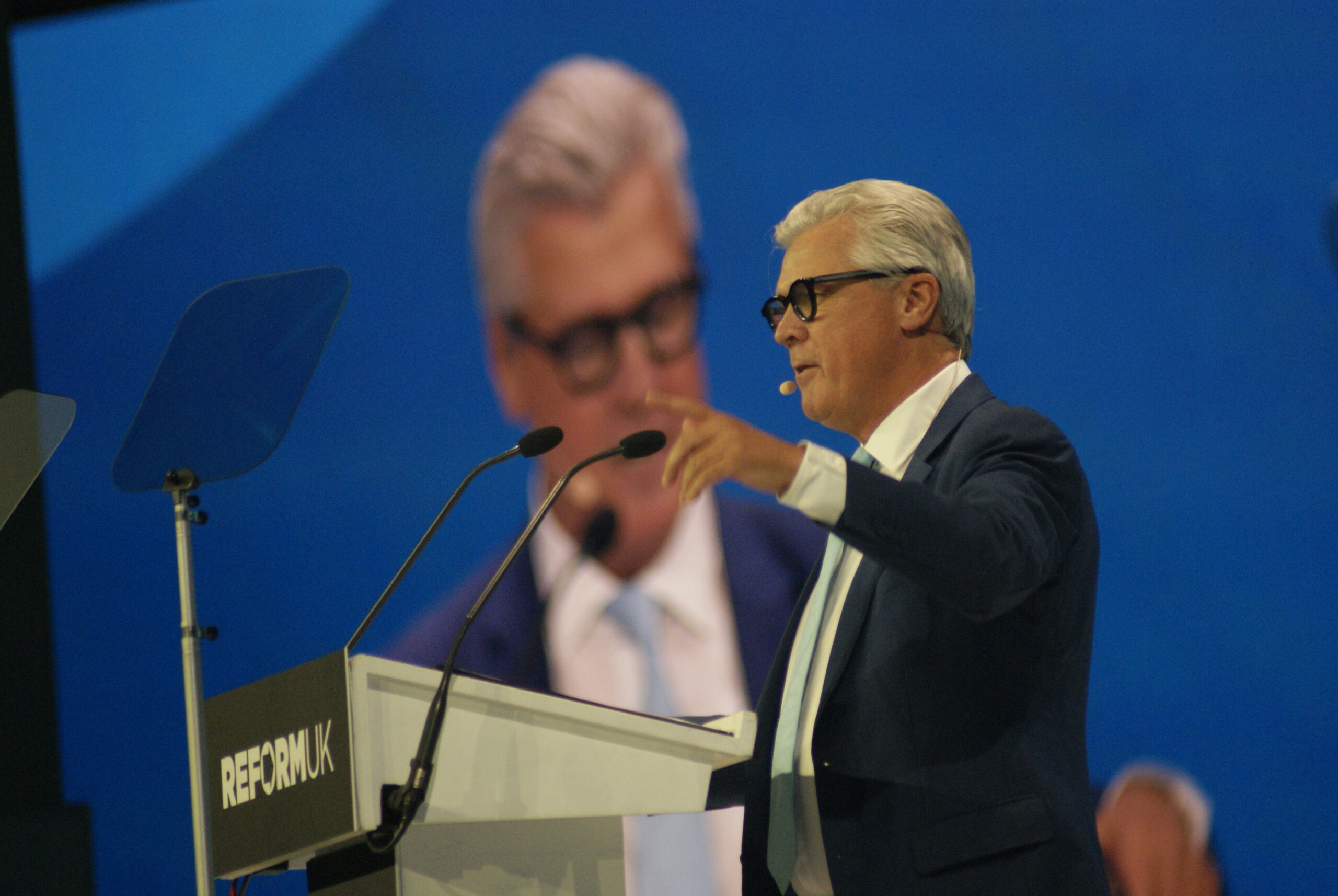 Malcolm Offord speaks from a Reform UK podium at conference, gesturing to the hall beneath a large screen showing his portrait.
