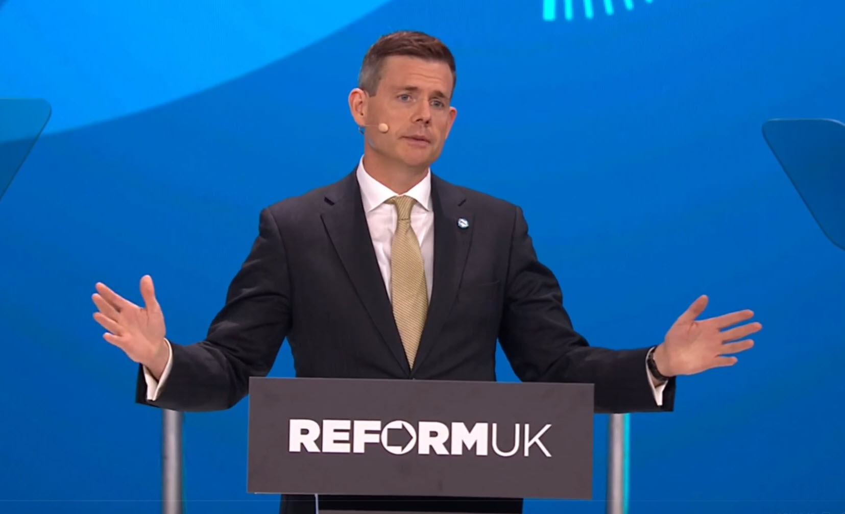 Matt Goodwin speaks at Reform UK Conference 2026 in Birmingham, gesturing from the Reform UK podium in a dark suit and gold tie.