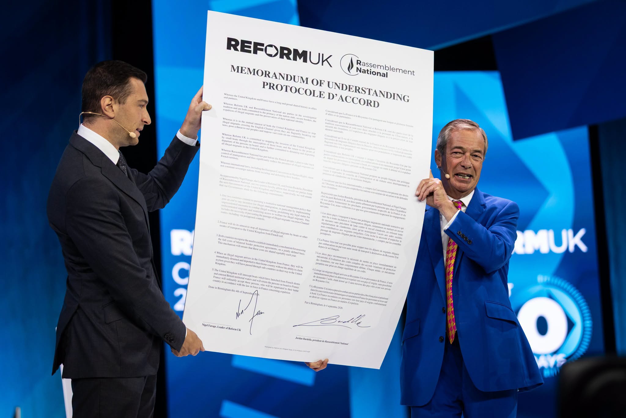Nigel Farage and Jordan Bardella on stage at Reform UK Conference 2026 in Birmingham, holding the signed bilingual Memorandum of Understanding between Reform UK and Rassemblement National.