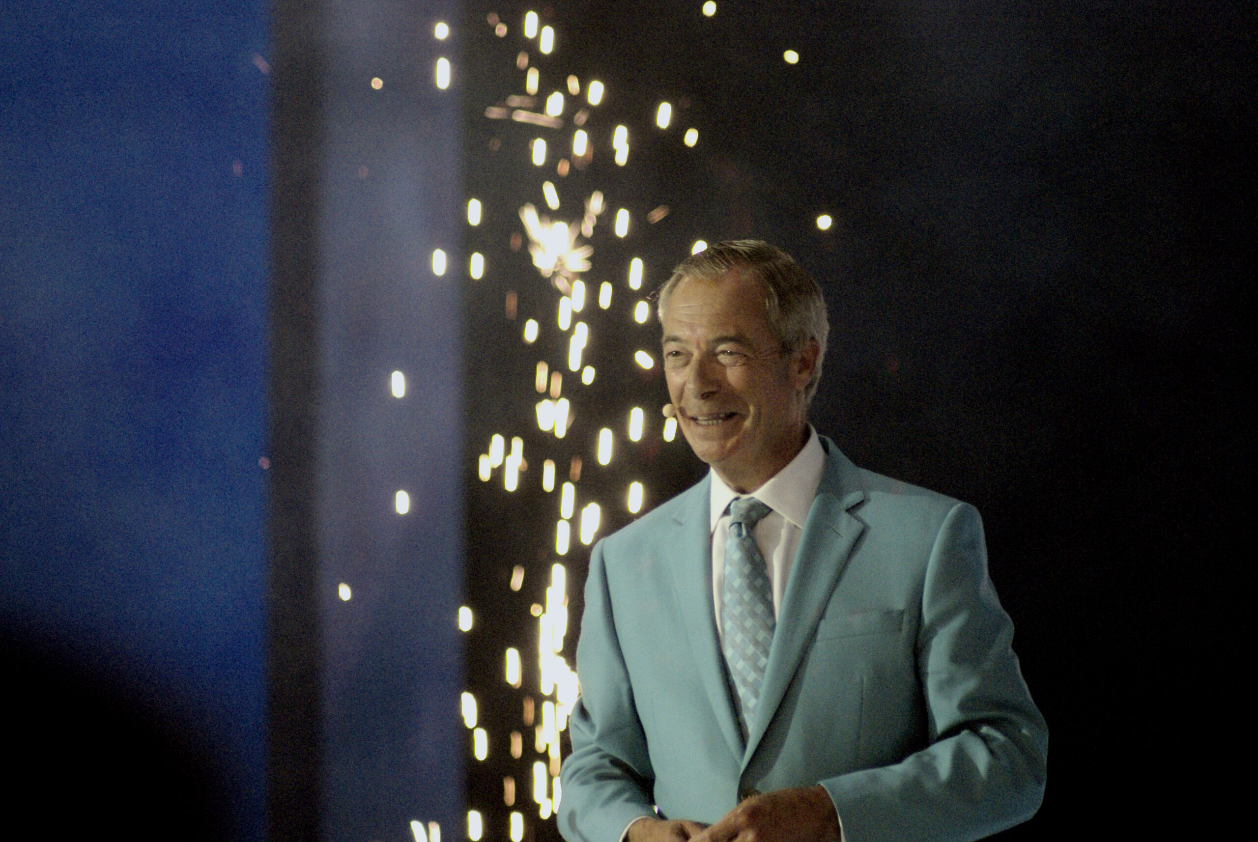 Nigel Farage closes Reform UK Conference 2026 from the podium in Birmingham, wearing a turquoise suit beneath a large on-stage portrait.