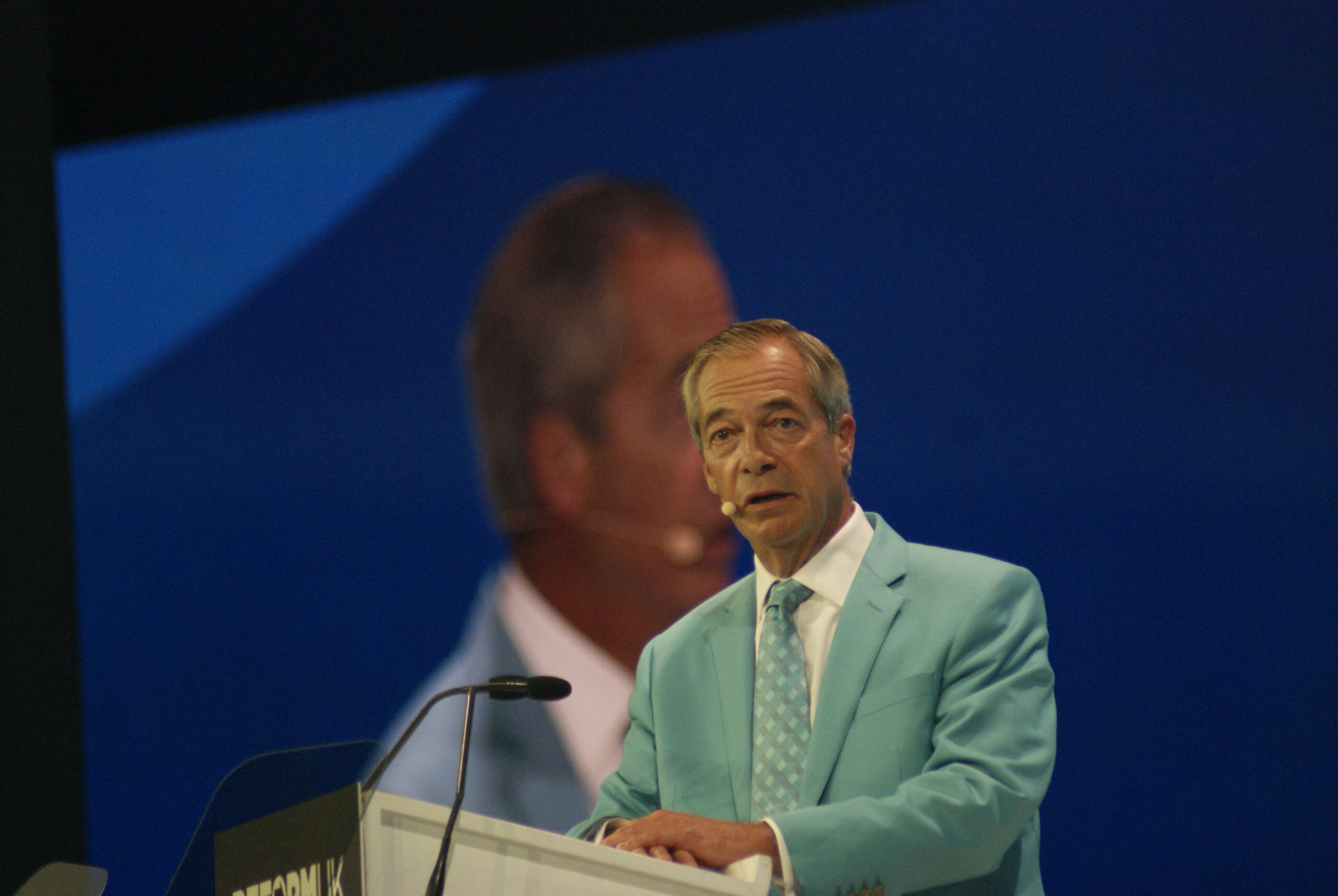 Nigel Farage on stage at Reform UK Conference 2026 as golden stage pyrotechnics erupt behind him.