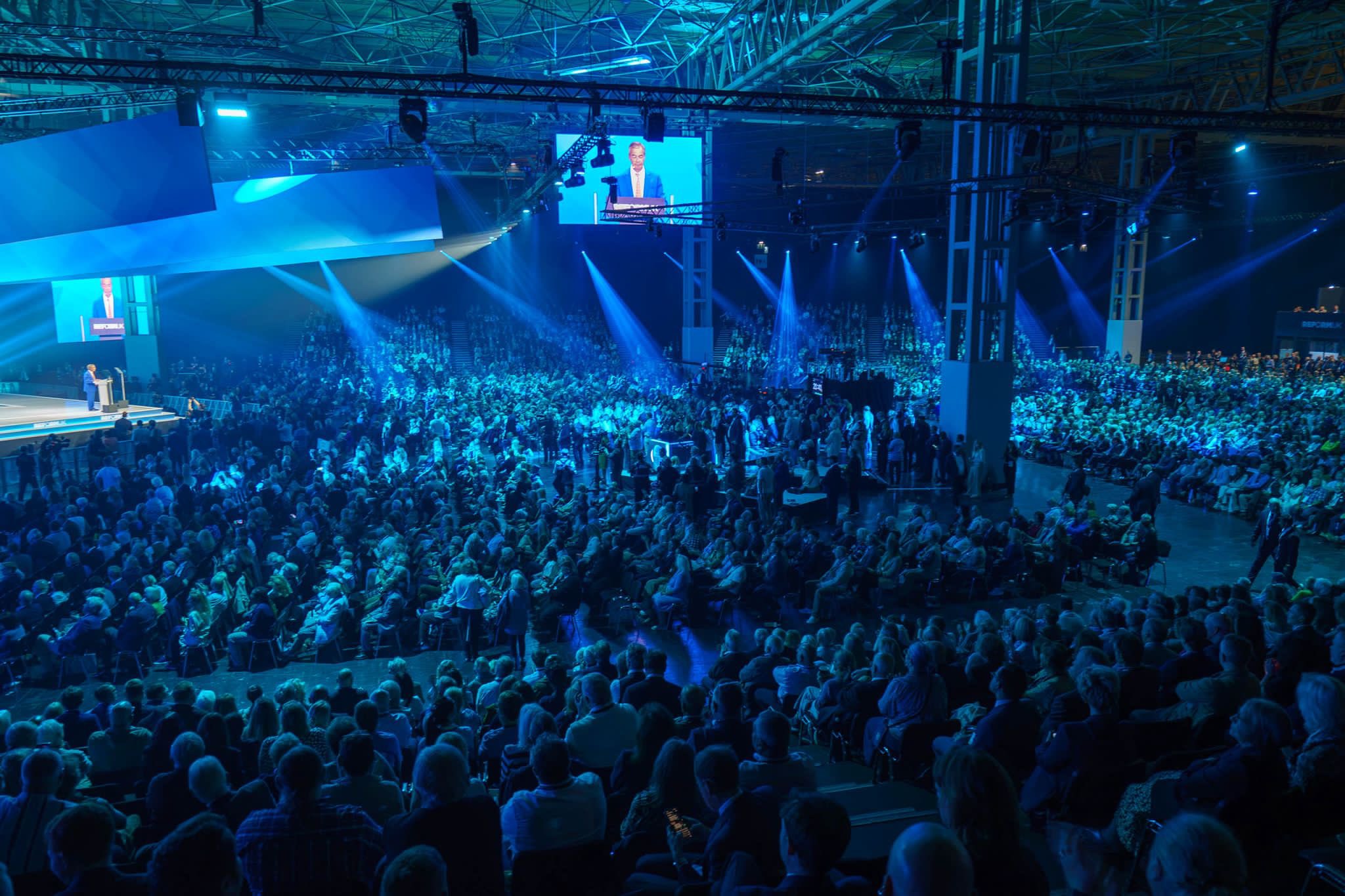 A wide view of Reform UK Conference 2026 at the Birmingham NEC, showing thousands of delegates facing the main stage under blue lighting and large screens.