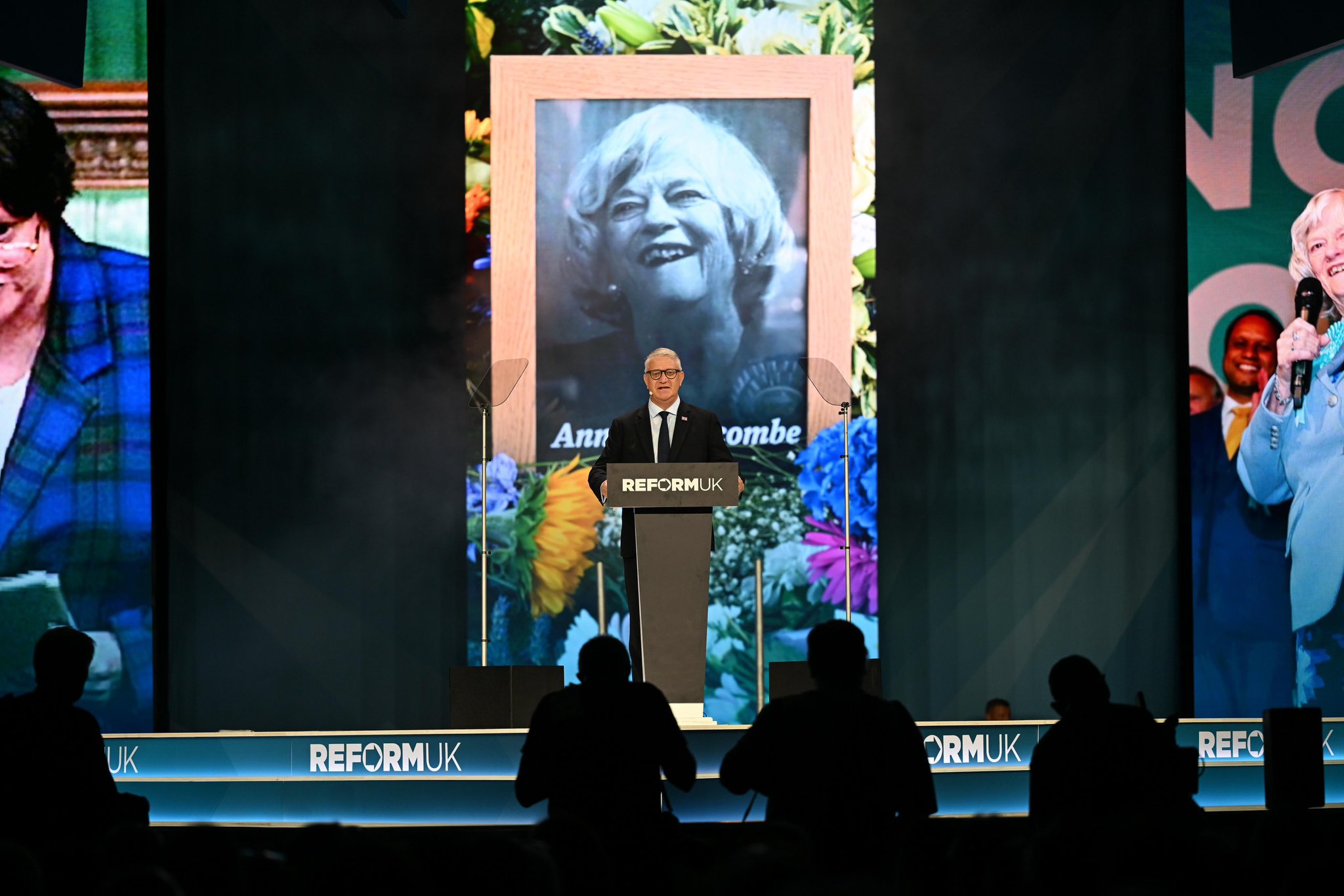 Nigel Farage stands at the Reform UK Conference podium beneath a large portrait of Ann Widdecombe during the opening tribute in Birmingham.