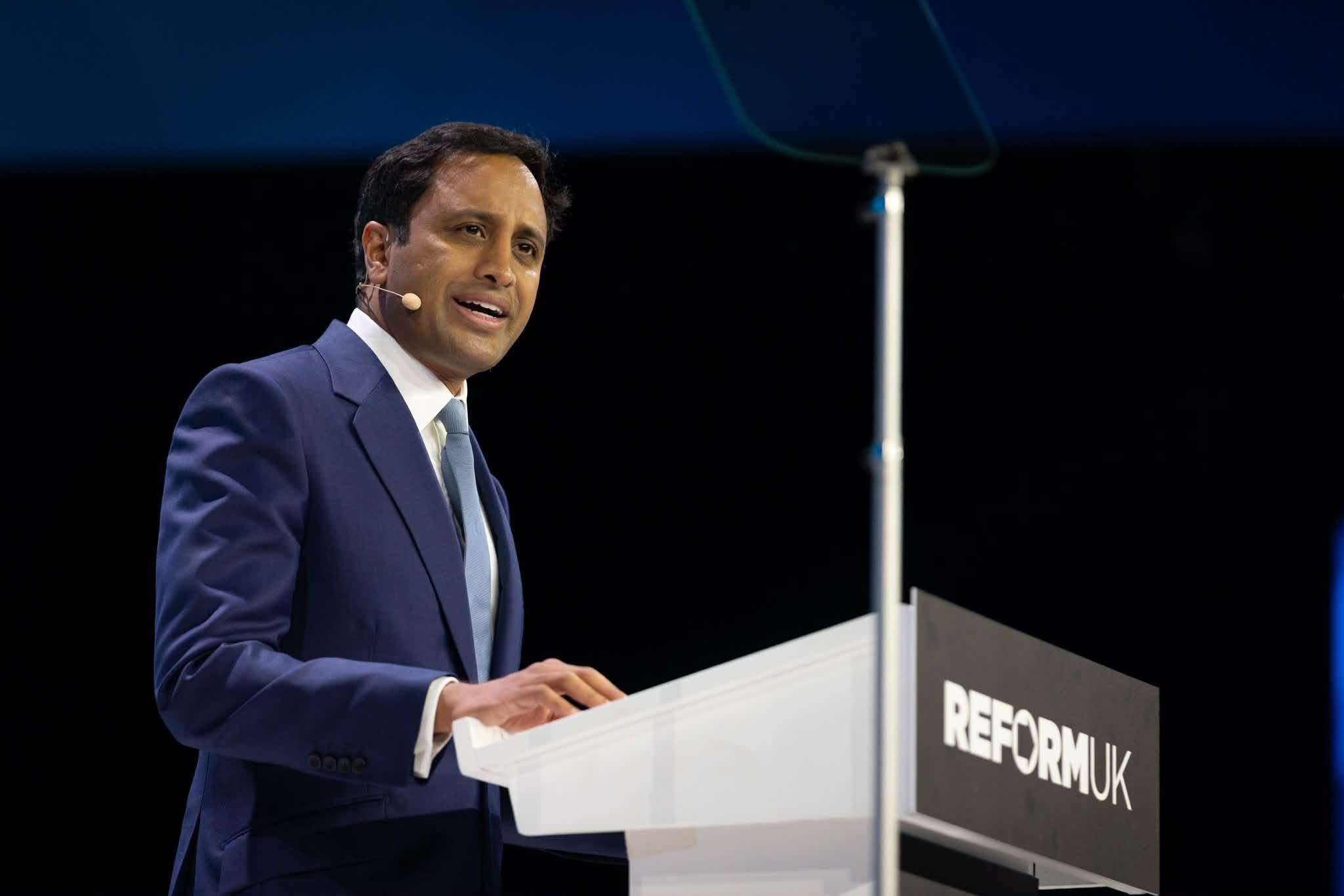 Zia Yusuf speaks at Reform UK Conference 2026 in Birmingham, standing at the Reform UK podium in a navy suit and light blue tie.